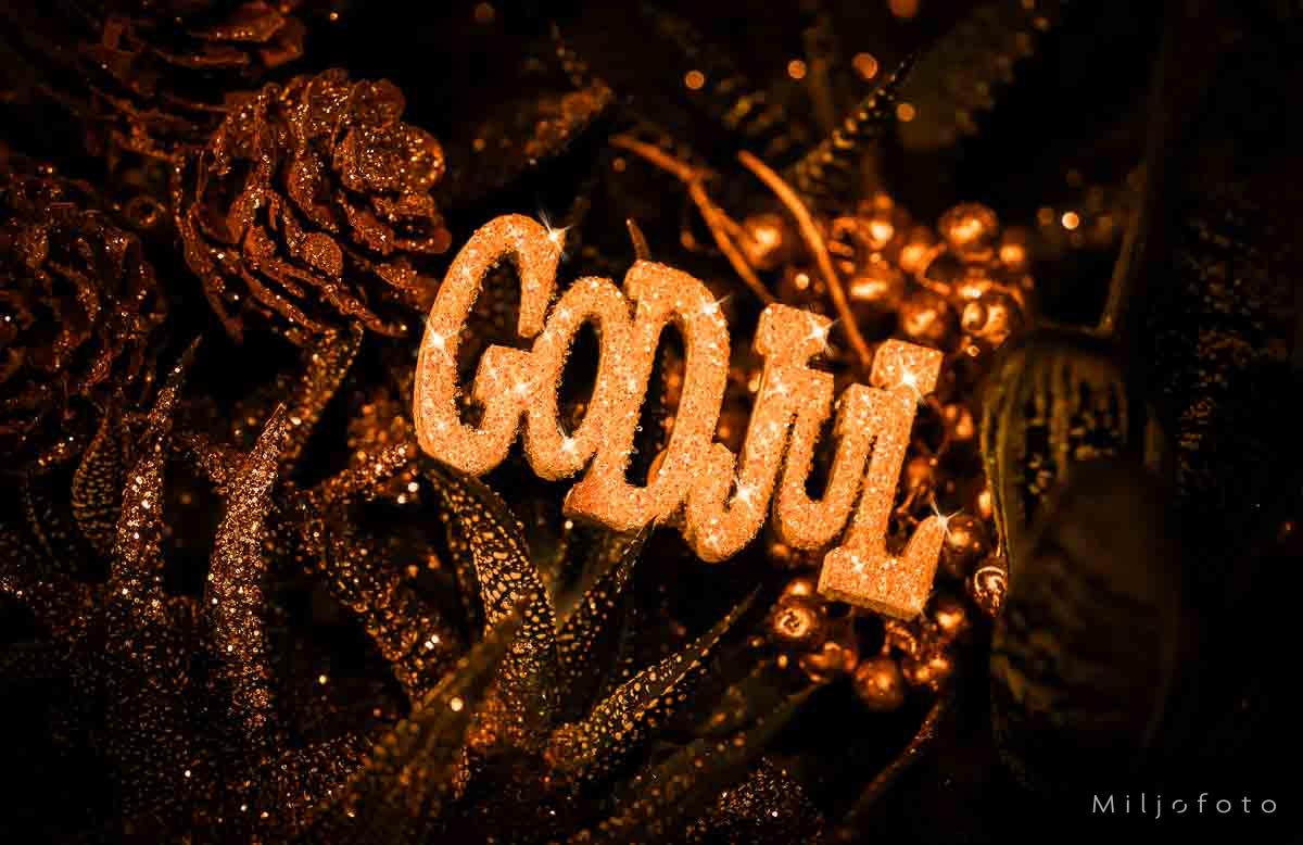 God jul i gull "Stemningsfullt bilde med teksten 'God Jul' i glitrende gull, omgitt av dekorative kongler og planter, med varme gyldne toner som skaper en koselig juleatmosfære."