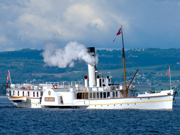 Skibladner, mjøsas hvite svane som seiler på en sommerblå Mjøsa, damp kommer opp av pipa.