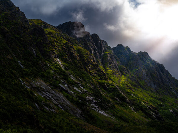 Bildet viser en bratt og dramatisk fjellside i Lofoten, preget av mørke bergvegger og frodig, grønn vegetasjon som klamrer seg til skråningene.