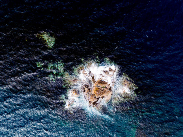 Bildet viser et lite, steinete skjær i havet fotografert ovenfra, der hvitt bølgeskum bryter mot steinene. Det dype, mørkeblå vannet omgir skjæret