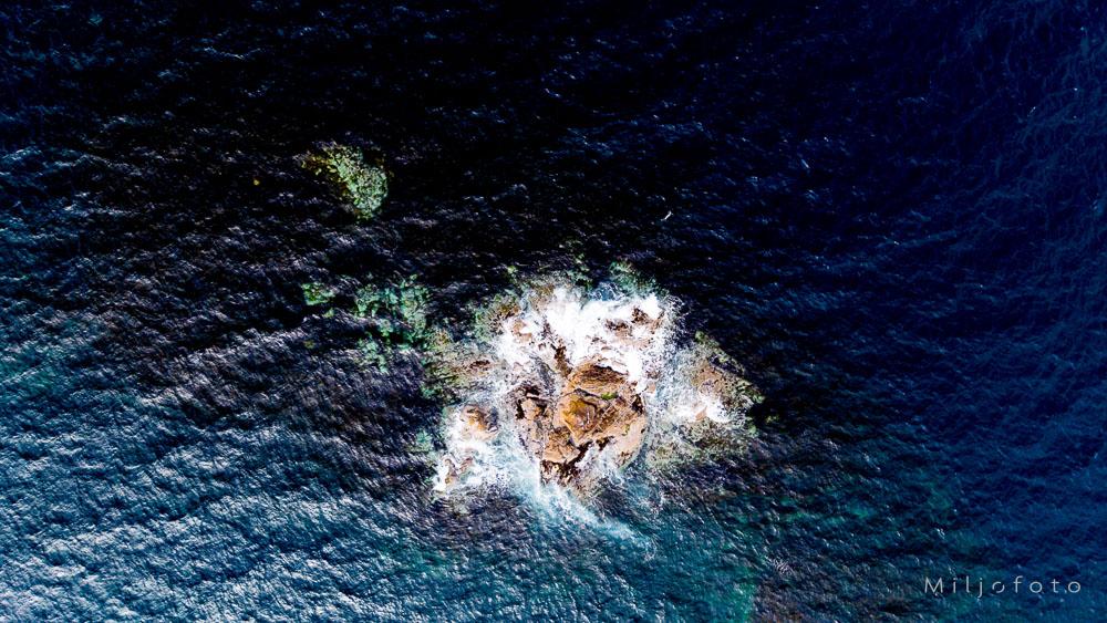 Bildet viser et lite, steinete skjær i havet fotografert ovenfra, der hvitt bølgeskum bryter mot steinene. Det dype, mørkeblå vannet omgir skjæret