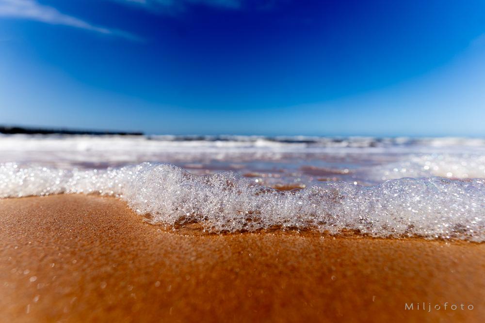 Bildet viser et nærbilde av glitrende, hvitt havskum som skyller opp på en gyllen sandstrand under en klar, dypblå himmel.
