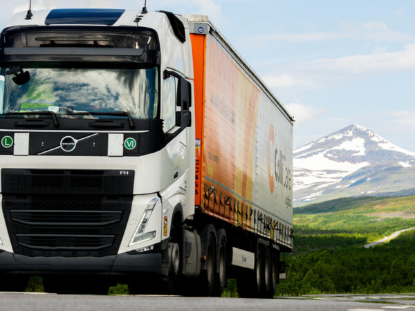 En hvit Volvo FH-lastebil med en oransje og hvit tilhenger kjører langs en vei i et vakkert naturlandskap.