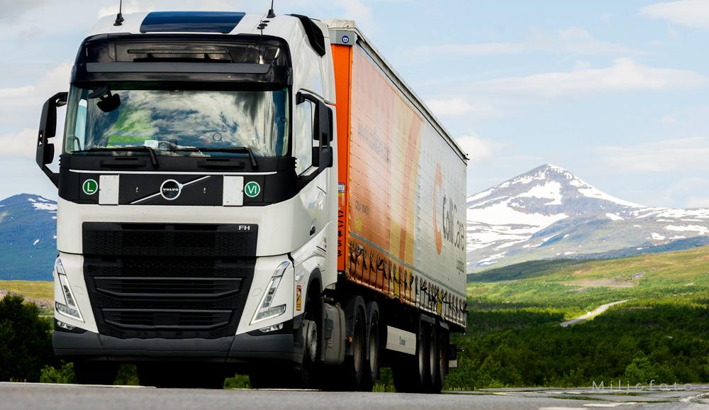 En hvit Volvo FH-lastebil med en oransje og hvit tilhenger kjører langs en vei i et vakkert naturlandskap.