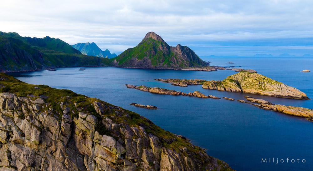 Bildet viser et vakkert og dramatisk kystlandskap fra Lofoten, med spisse, grønnkledde fjell som reiser seg opp fra det dype